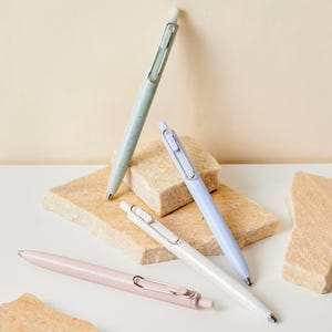 May include: Four pastel-coloured pens: light green, light blue, white, and pink. Each pen has a silver clip and a speckled design. The pens are arranged on a white surface with beige stone blocks.