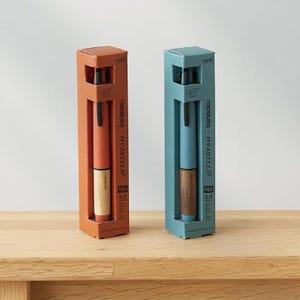 May include: Two pens in boxes. One pen is orange with a wooden grip, the other is blue with a wooden grip. The boxes are labeled "JETSTREAM x karimoku" and have the text "0.5". The pens are on a wooden surface.