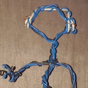 May include: A wire sculpture of a person made from blue and black electrical wire. The sculpture is posed with arms outstretched and a head tilted to the side.