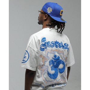 Könnte beinhalten: Hellgraues T-Shirt mit einem großen blauen Om-Symbol und dem Wort "Bestes" in blauer Schrift. Das Shirt hat auch die Worte "Reach Inner Peace". Eine blaue Baseballkappe mit einem gelben "C" wird getragen.