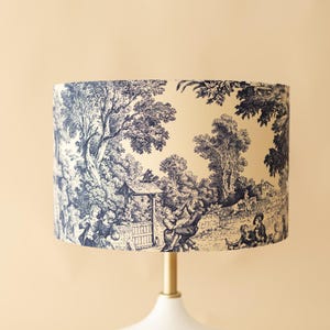 Pode incluir: Um abajur cilíndrico com um padrão toile azul-escuro sobre um fundo creme. O design intrincado retrata cenas pastorais com árvores, folhagem e figuras com roupas de época. O abajur está montado sobre uma haste de metal dourado.