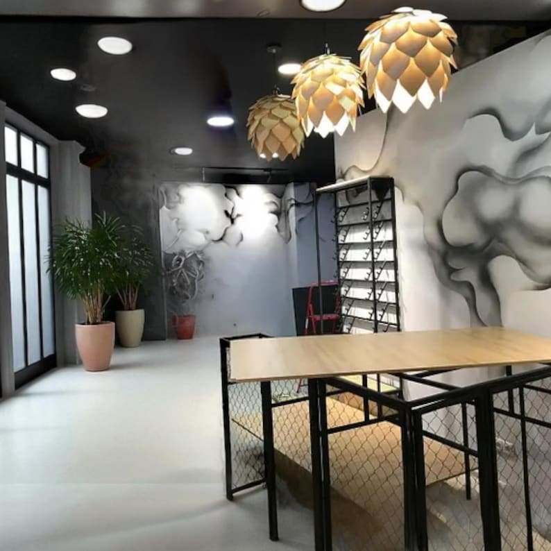 K&ouml;nnte beinhalten: Innenaufnahme eines modernen Gesch&auml;fts mit einem hellen Holztisch und einem schwarzen Metallrahmen. Drei dekorative Pendelleuchten h&auml;ngen von der schwarzen Decke. Die W&auml;nde sind mit einem grau-wei&szlig;en abstrakten Design bemalt. Topfpflanzen stehen in der Ecke.