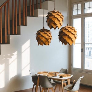 K&ouml;nnte beinhalten: Drei Pendelleuchten in Kiefernzapfenform h&auml;ngen &uuml;ber einem Esstisch. Die Leuchten sind aus hellem Holz gefertigt. Der Tisch ist wei&szlig; mit einem hellen Holzsockel, umgeben von grauen St&uuml;hlen. Im Hintergrund sind eine Treppe und ein Fenster zu sehen.