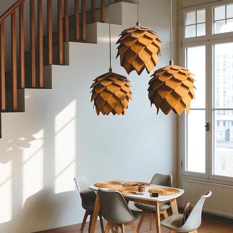 K&ouml;nnte beinhalten: Drei Pendelleuchten in Kiefernzapfenform h&auml;ngen &uuml;ber einem Esstisch. Die Leuchten sind aus hellem Holz gefertigt. Der Tisch ist wei&szlig; mit einem hellen Holzsockel, umgeben von grauen St&uuml;hlen. Im Hintergrund sind eine Treppe und ein Fenster zu sehen.