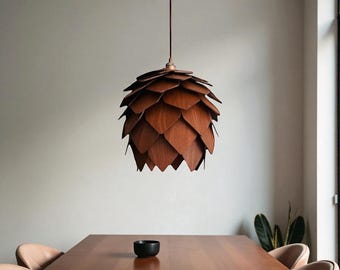 Op de afbeelding: Een houten hanglamp in de vorm van een dennenappel, opgehangen aan een bruin snoer. De lamp is gemaakt van gelaagde, donkerbruine houten panelen. De lamp hangt boven een houten eettafel.