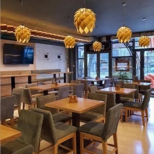 K&ouml;nnte beinhalten: Innenansicht eines Restaurants mit Holztischen und St&uuml;hlen. Die Decke ist mit mehreren kegelf&ouml;rmigen Pendelleuchten ausgestattet. Ein gro&szlig;er Fernseher ist an der Wand montiert. Der Raum hat gro&szlig;e Fenster und ein modernes Design.