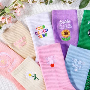 Könnte beinhalten: Verschiedene bunte Socken mit gestickten Designs und Texten. Zu den Designs gehören eine Sonnenblume, ein Fußball und die Wörter "Best Mom", "Pilates Pals" und "Your Design Here". Die Socken sind auf einer weißen, strukturierten Oberfläche angeordnet.