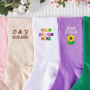 Könnte beinhalten: Fünf Paar bunte Socken mit gestickten Designs. Die Socken sind rosa, beige, weiß, lila und grün. Texte sind "BEST MOM" mit Herz, "O & Y 01.01.2026", "YOUR DESIGN HERE", "Bride 10.10.26" mit Sonnenblume und "PLAYER NO.1" mit Fußball.