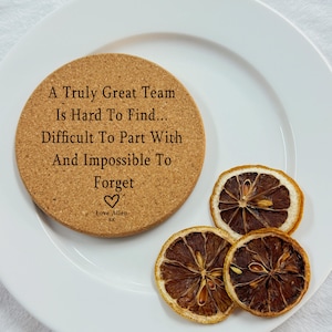 May include: A round cork coaster with the text "A Truly Great Team Is Hard To Find... Difficult To Part With And Impossible To Forget." The coaster is on a white plate with three dried lemon slices.