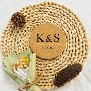 Puede incluir: Un posavasos redondo de corcho con las iniciales "K&S" y la fecha "05.22.2023" impresas en negro. El posavasos est&aacute; sobre una estera de paja tejida.