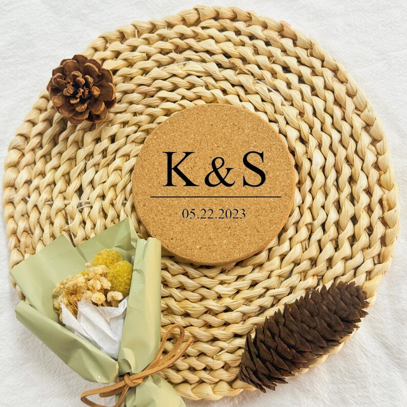 Puede incluir: Un posavasos redondo de corcho con las iniciales "K&S" y la fecha "05.22.2023" impresas en negro. El posavasos est&aacute; sobre una estera de paja tejida.