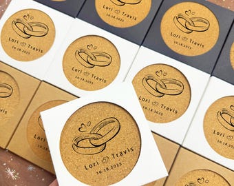 May include: Cork coasters with a wedding ring design and the names "Lori & Travis" along with the date "10.18.2025" printed on them. The coasters are in white, black, and brown boxes, ideal for wedding favors.