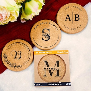 Könnte beinhalten: Eine Sammlung personalisierter Korkuntersetzer. Jeder Untersetzer zeigt individuelle Texte und Designs, darunter Initialen, Namen und Daten. Ein Untersetzer ist in einer Pappschachtel mit dem Text "Thank You" verpackt.