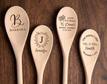 Può includere: Quattro cucchiai in legno chiaro con testi e disegni incisi al laser. Il primo presenta una grande lettera B e il nome BARBARA. Il secondo mostra una lettera J all'interno di uno stemma floreale con il nome Jennifer. Il terzo mostra un bouquet floreale con il testo LOVE is in bloom Eliana's BRIDAL SHOWER 12.05.2026. Il quarto riporta THE PERFECT MIX Mr. & Mrs. Smith DECEMBER 5, 2026.