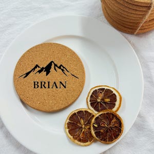 Puede incluir: Un posavasos redondo de corcho con una silueta de monta&ntilde;a negra y el nombre "BRIAN" impreso en letras negras.