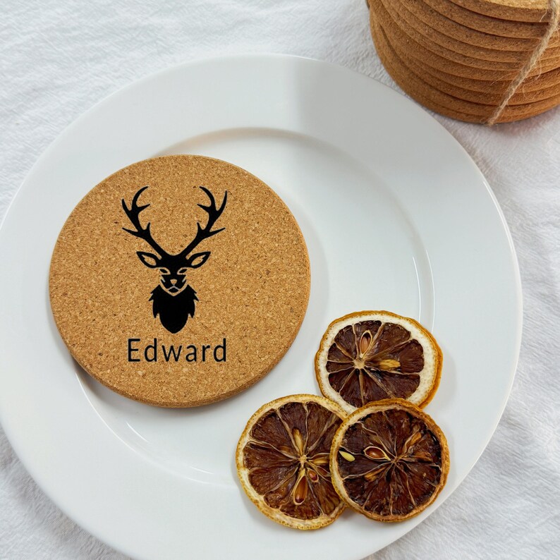 Puede incluir: Un posavasos redondo de corcho con una silueta negra de un ciervo con cuernos y el nombre "Edward" impreso debajo. El posavasos est&aacute; sobre un plato blanco con rodajas de lim&oacute;n secas.
