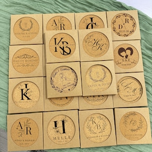 Puede incluir: Un conjunto de 12 posavasos de corcho personalizados con iniciales y nombres grabados. Cada posavasos está en una caja cuadrada con fondo marrón. Los posavasos presentan una variedad de diseños, incluyendo coronas de flores, corazones y caligrafía.