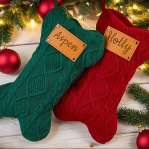 以下が含まれることがあります： ケーブル編みのデザインの、緑と赤のクリスマスストッキングが2つ。各ストッキングには、名前が書かれた茶色の革タグが付いています。「Aspen」と「Holly」です。ストッキングはクリスマスの飾りと緑で飾られています。