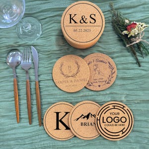 Puede incluir: Seis posavasos redondos de corcho con diferentes dise&ntilde;os. Los posavasos presentan texto personalizado, incluidos nombres, fechas y un logotipo. Los posavasos est&aacute;n sobre una superficie de tela verde.