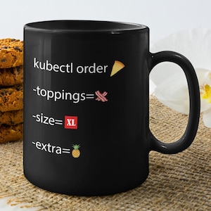 May include: Black coffee mug with white text that reads "kubectl order -toppings=🥓 -size=XL -extra=🍍". The mug is sitting on a brown burlap surface.