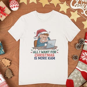 May include: White t-shirt with a humorous Christmas graphic. The design features a stressed cartoon person wearing a Santa hat, working on a laptop, with the text "ALL I WANT FOR CHRISTMAS IS MORE RAM".