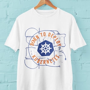 May include: White t-shirt with a blue and orange graphic design featuring a stylized image of a ship's wheel and the text "Born to Deploy Kubernetes".