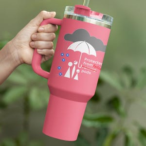 May include: Pink stainless steel travel mug with a handle and a straw. The mug features a graphic of a person under an umbrella with the text "Protected from falling training pods".