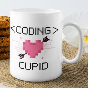 Op de afbeelding: Witte keramische mok met een roze gepixelde hart met een pijl erdoorheen. De mok heeft de tekst "<CODING> CUPID" erop gedrukt.