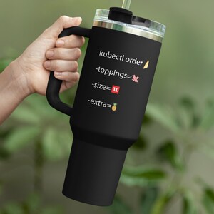May include: Black insulated tumbler with a handle, clear lid, and straw. The tumbler has text that reads "kubectl order -toppings=X -size= XL -extra=🍍". The background is a blurred green.