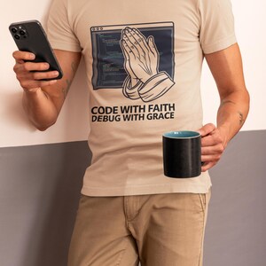 Puede incluir: Una camiseta beige de manga corta con un gráfico de manos en oración superpuesto sobre una ventana de código informático. El texto "CODE WITH FAITH DEBUG WITH GRACE" está impreso debajo del gráfico. La camiseta se combina con pantalones color caqui.