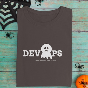 May include: Dark gray crew neck t-shirt with the word "DEVOPS" and a ghost graphic in white. The text "WHERE MONSTERS COME TO LIFE" is below the graphic. Halloween-themed apparel.