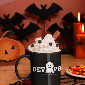 Könnte beinhalten: Schwarze Tasse mit dem Wort "DEVOPS" und einer Geistergrafik, gefüllt mit Schlagsahne, Schokoladenstückchen und einem Zimtstange. Die Tasse steht auf einem roten Untersetzer, mit Halloween-Dekorationen im Hintergrund.