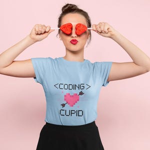 Op de afbeelding: Een vrouw draagt een lichtblauw T-shirt met de tekst "<CODING> CUPID" en een gepixelde hart met een pijl erdoorheen. Ze houdt twee rode hartvormige lolly's voor haar ogen.