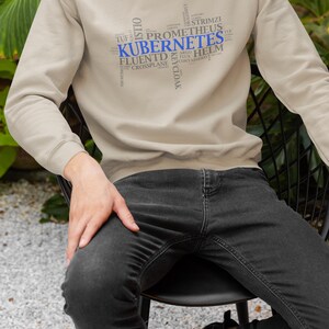 Puede incluir: Una sudadera beige con un diseño de nube de palabras en azul y gris con la palabra "Kubernetes". La sudadera la lleva puesta una persona sentada en una silla negra.
