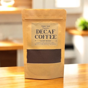 Könnte beinhalten: Ein brauner Papiersack mit entkoffeiniertem Kaffee und einem rechteckigen Fenster, das das dunkle Kaffeepulver zeigt. Der Beutel hat ein Etikett mit dem Text "DECAF COFFEE" und "LIGHT ROAST". Das Etikett gibt auch an, dass es sich um Bio-Arabica-Kaffeepulver handelt.
