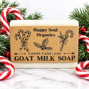 Puede incluir: Una pastilla rectangular de jabón de leche de cabra blanco envuelta en una etiqueta de papel color canela. La etiqueta presenta ilustraciones en tinta negra de bastones de caramelo y acebo, con el texto Happy Soul Organics, Candy Cane, 113 g.
