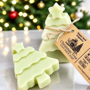 Puede incluir: Dos pastillas de jabón de leche de cabra de color verde claro con forma de árbol de Navidad. Una pastilla está en posición vertical, atada con cordel a una etiqueta de papel marrón que dice Happy Soul Organics, Christmas Tree GOAT MILK SOAP.