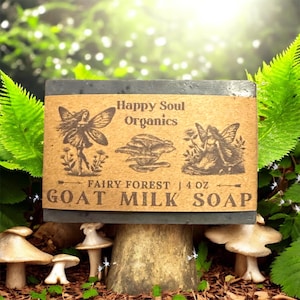 May include: A dark gray goat milk soap bar with a brown label featuring fairy illustrations and the text "Happy Soul Organics Fairy Forest | 4 oz GOAT MILK SOAP". The soap is displayed on a mushroom-shaped stand, surrounded by greenery.