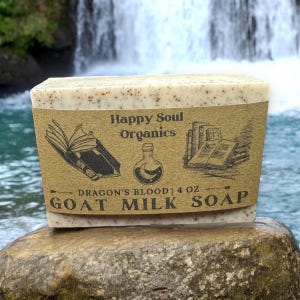 May include: A bar of goat milk soap with a brown paper label that reads "Happy Soul Organics" and "Dragon's Blood | 4 oz Goat Milk Soap". The label features illustrations of books and a potion bottle.