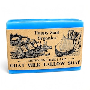 May include: A rectangular bar of bright blue soap wrapped in a tan paper label. The label features black ink illustrations of a Viking village and a longship, with the text Happy Soul Organics, Methylene Blue, 4 oz, and Goat Milk Tallow Soap.