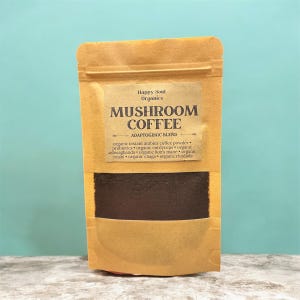 以下が含まれることがあります： 「Happy Soul Organics」と「Adaptogenic Blend」の文字が入った「Mushroom Coffee」のベージュ色のポーチ。オーガニックインスタントアラビカコーヒーパウダー、プロバイオティクス、オーガニックライオンズメインなどの成分が記載されています。ポーチには濃い茶色のコーヒーが入っています。