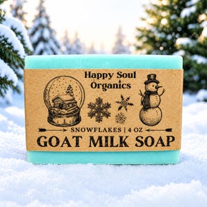 Puede incluir: Una pastilla rectangular de jabón de leche de cabra azul claro envuelta en una etiqueta de papel marrón. La etiqueta incluye el texto Happy Soul Organics, SNOWFLAKES | 4 OZ, GOAT MILK SOAP, junto con ilustraciones de una bola de nieve, copos y un muñeco.