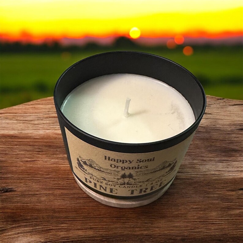 Puede incluir: Un recipiente negro para velas que contiene cera blanca con una mecha. La etiqueta presenta el texto Happy Soul Organics, PURE SOY CANDLE 4 OZ y PINE TREE, acompa&ntilde;ado de una ilustraci&oacute;n de un paisaje con monta&ntilde;as y &aacute;rboles sobre un fondo beige.