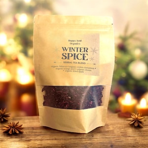Pode incluir: Um saco de papel kraft de mistura de chá de ervas "Winter Spice". O saco tem uma janela transparente mostrando as folhas de chá. O rótulo lista os ingredientes: hibisco orgânico, canela, casca de laranja, cravinho e maçã seca. O fundo inclui velas e anis estrelado.
