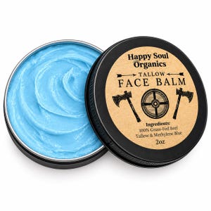 Op de afbeelding: Een ronde zwarte metalen blik met felblauwe gezichtsbalsem, met het deksel ernaast geplaatst. Het deksel heeft een beige label met de tekst Happy Soul Organics, TALLOW FACE BALM, en ingrediënten 100% Grass-Fed Beef Tallow & Methylene Blue 57g.