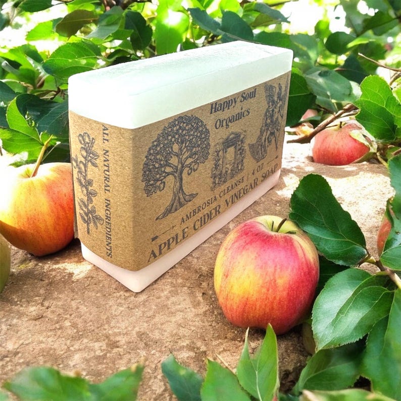 May include: A rectangular bar of soap with a white top and a brown label featuring a tree illustration and the text "Happy Soul Organics Apple Cider Vinegar Soap". Red and yellow apples and green leaves surround the soap.