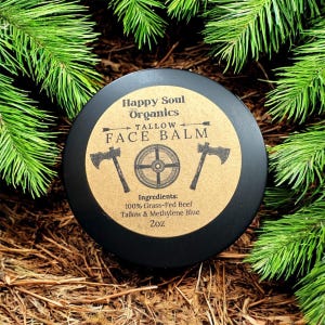 Może przedstawiać: Czarny słoik z brązową etykietą, na której widnieje napis "Happy Soul Organics Tallow Face Balm". Etykieta zawiera również tarczę w stylu wikingów i dwie siekiery. Składniki są wymienione jako "100% Grass-Fed Beef Tallow & Methylene Blue", a rozmiar to "2oz".