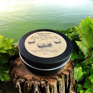 K&ouml;nnte beinhalten: Ein runder schwarzer Metallbeh&auml;lter mit Happy Soul Organics Natural Body Scrub. Das Etikett zeigt eine Illustration eines alten Schiffes und listet Inhaltsstoffe wie Meersalz, Rizinus&ouml;l, Vitamin E, Kokos&ouml;l und &auml;therische &Ouml;le auf.