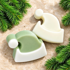 Puede incluir: Dos pastillas de jabón sólidas con forma de gorro de Papá Noel. Un jabón es totalmente blanco roto, mientras que el otro tiene el cuerpo del gorro en verde salvia con el borde y el pompón blancos. Los jabones están junto a ramas de pino.