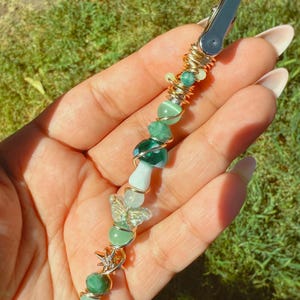 May include: A green and gold hair clip with a variety of beads, including a mushroom, butterfly, and star. The clip is made with wire and has a silver metal clasp.